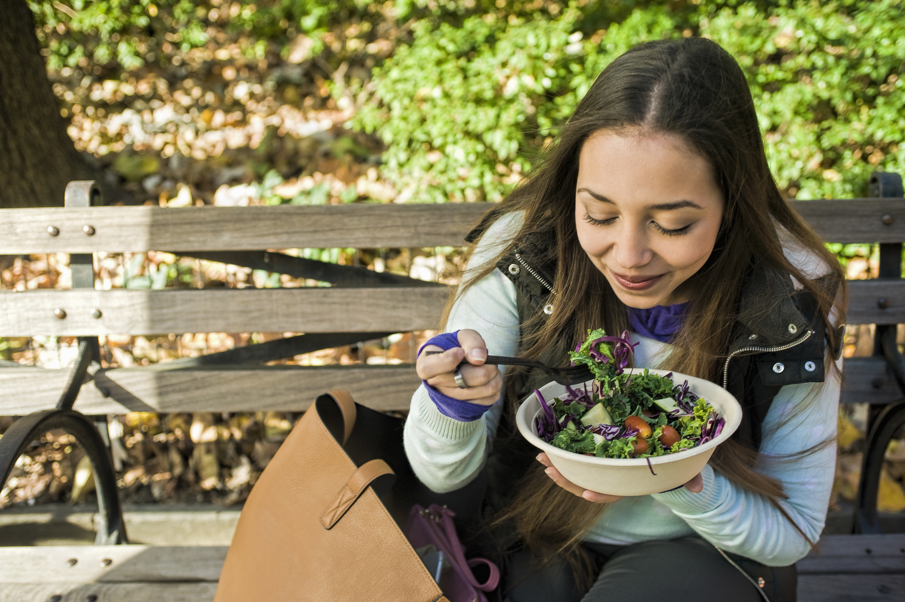 A woman eats a salad while sitting on a park bench