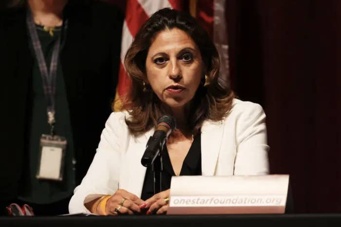 Mitchell sits at a press conference with a microphone wearing a white blazer