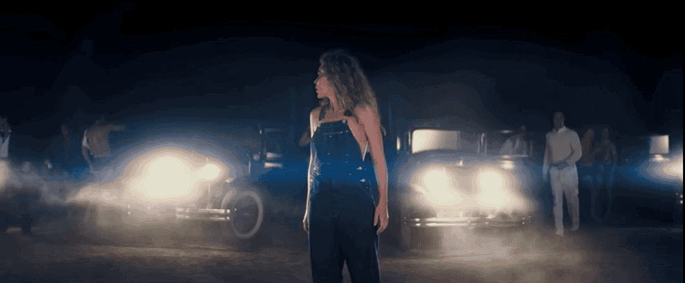 Movie gif: Margot Robbie wearing overalls in front of parked cars with headlights on