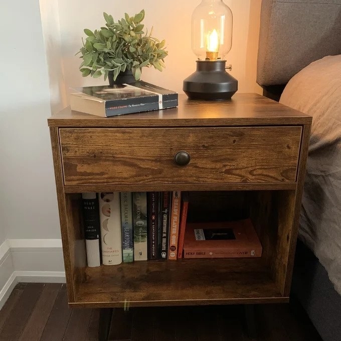 a reviewer photo of the wooden square shaped bed table with top drawer, books in bottom cubby and lamp, plant, and book on the top