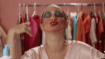gif of a person in makeup posing confidently among clothing racks, with a whimsical expression