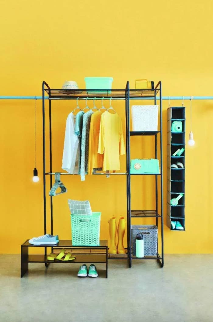 the black metal freestanding closet on a yellow background, filled with yellow and teal clothes and accessories