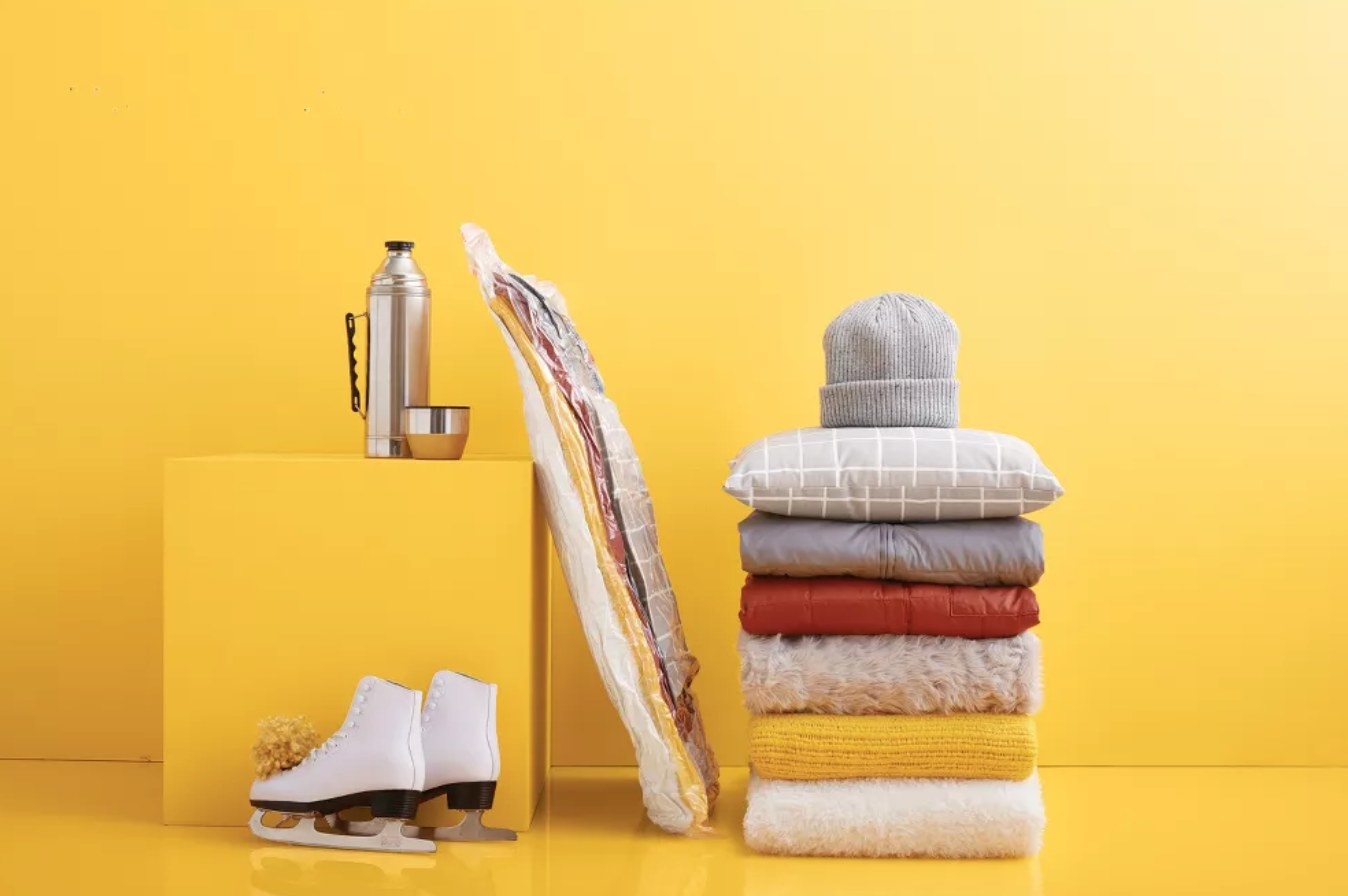 a set of winter clothes next to the same clothes in the vacuum bag, as well as ice skates and a winter thermos — all against a bright yellow background
