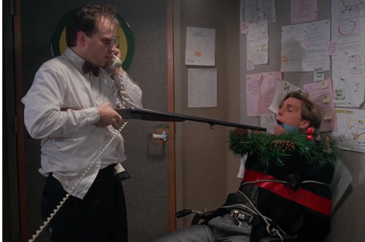 eliot holds a shotgun at a network employee who is tied up in tinsel