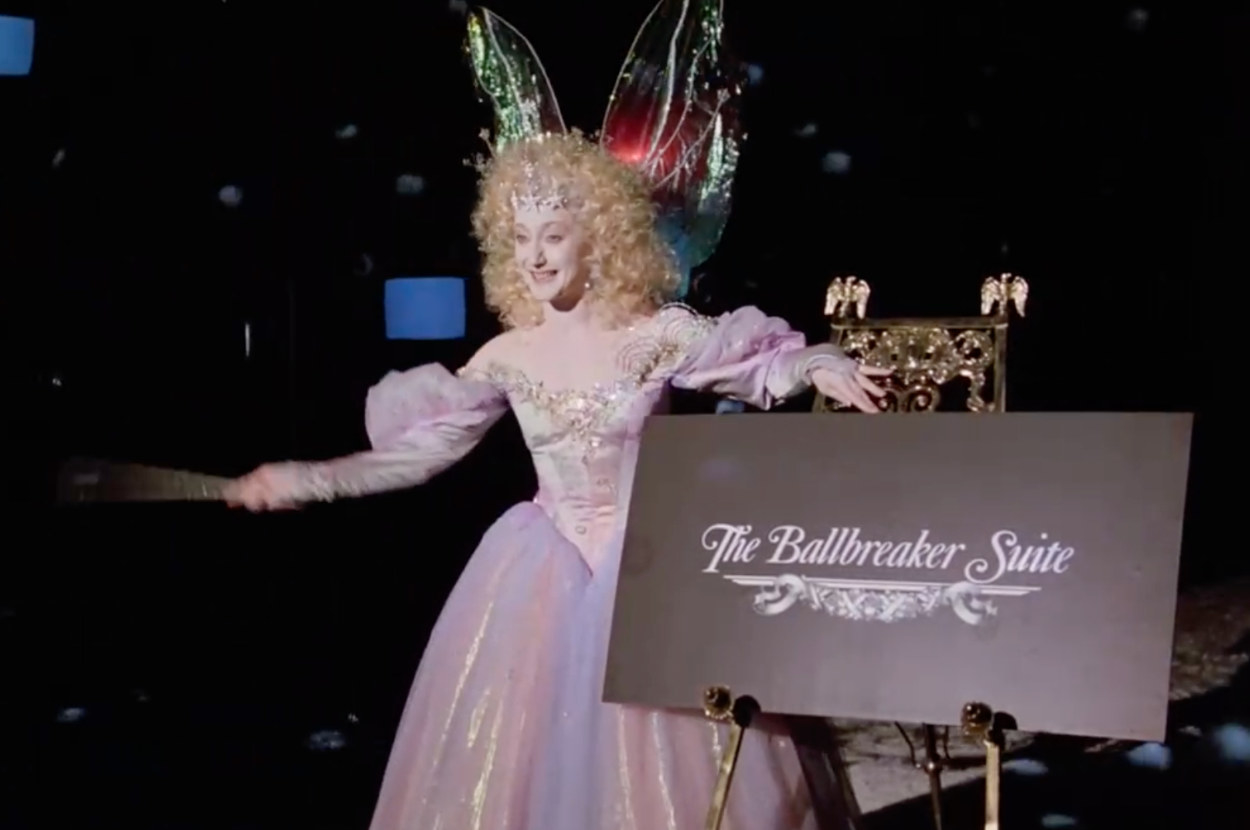 Carol Kane as the Ghost of Christmas Present stands next to a sign the reads the Ballbreaker Suite