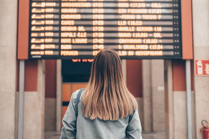 A young woman at a railway station or at the airport looks at the smartphone screen against the background of the arrival and departure board