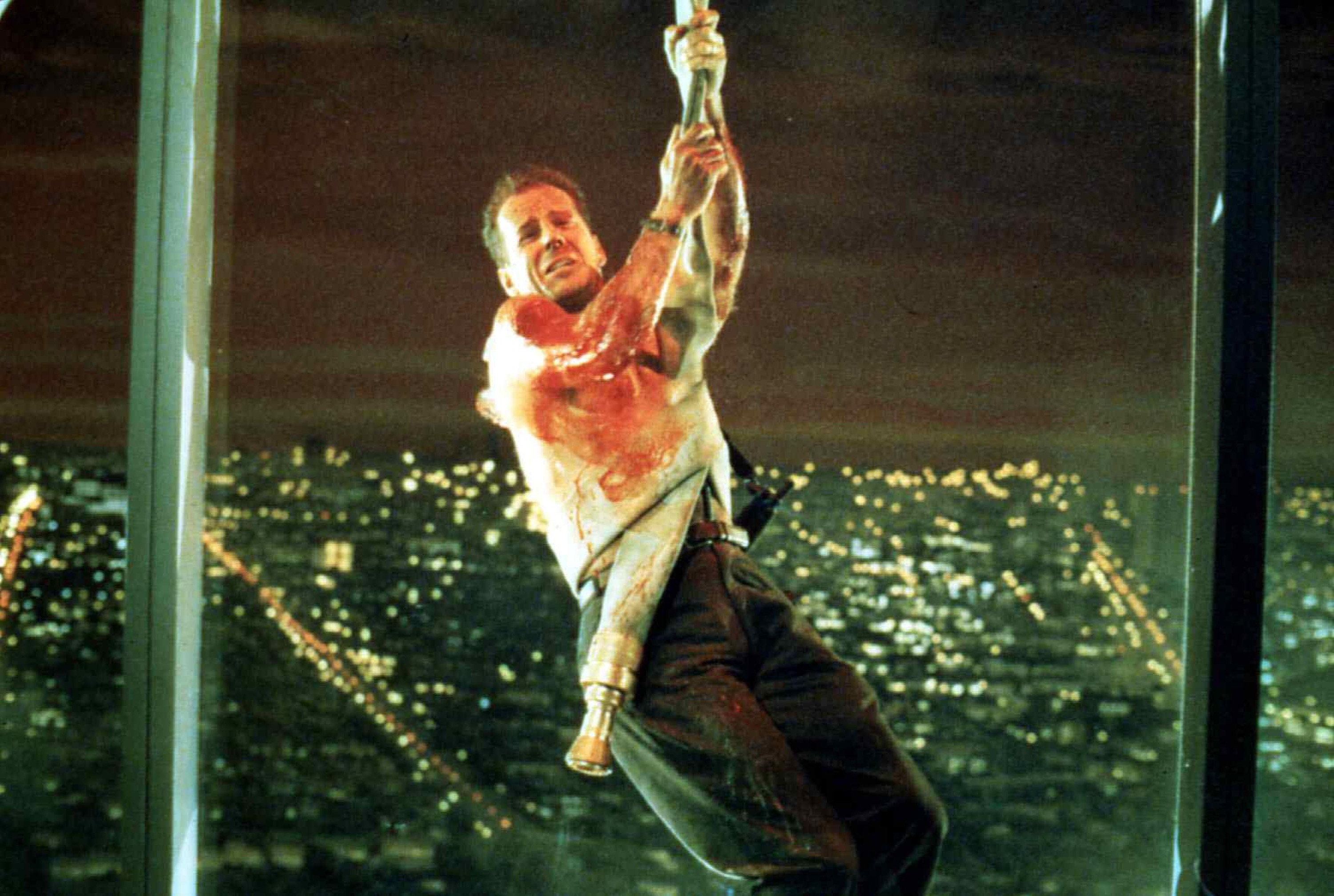 A bloodied man in a tanktop and jeans swings into a building window with a fire hose