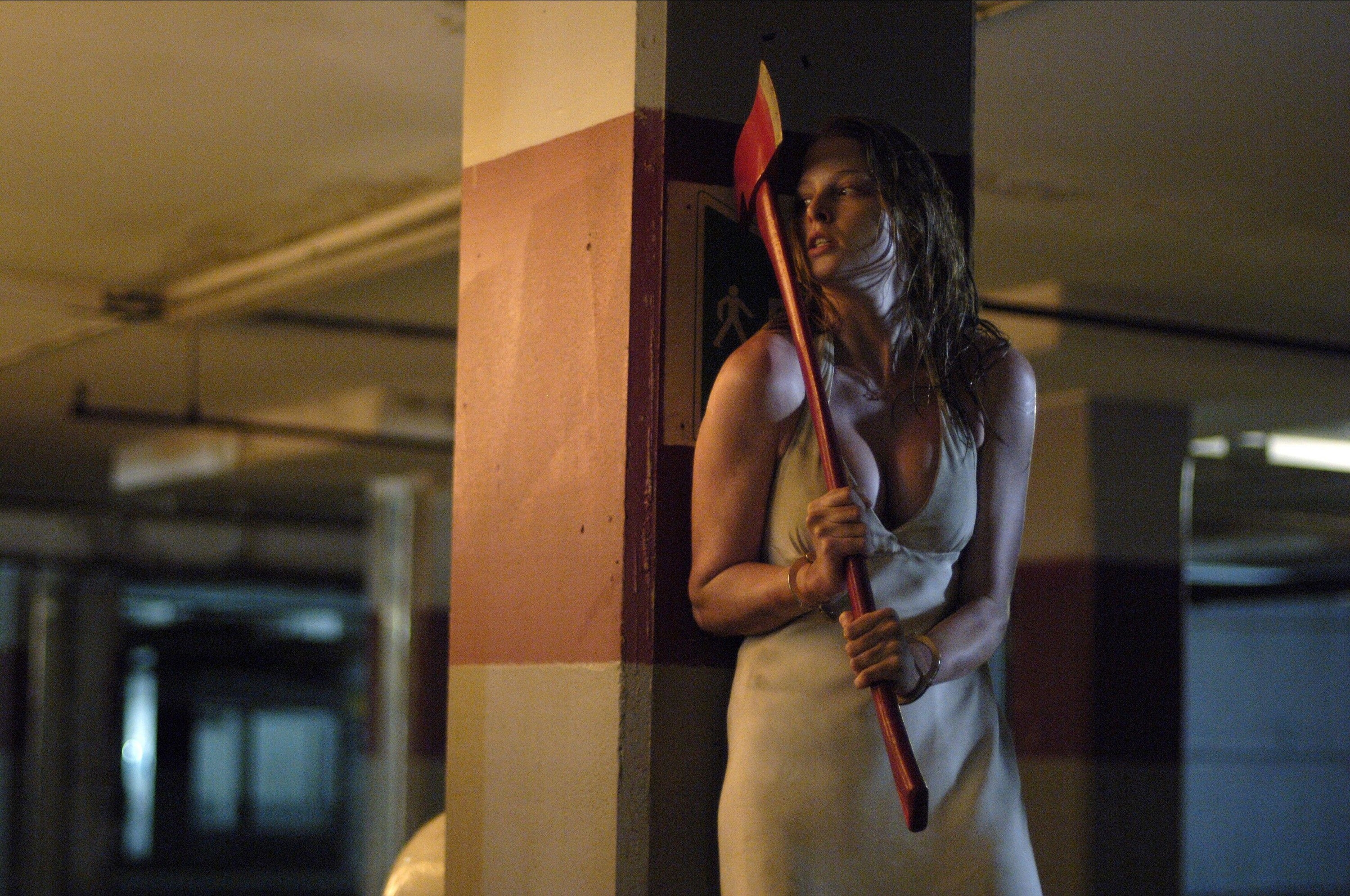 A woman in a revealing white dress hides near a pillar in a parking garage while holding an axe