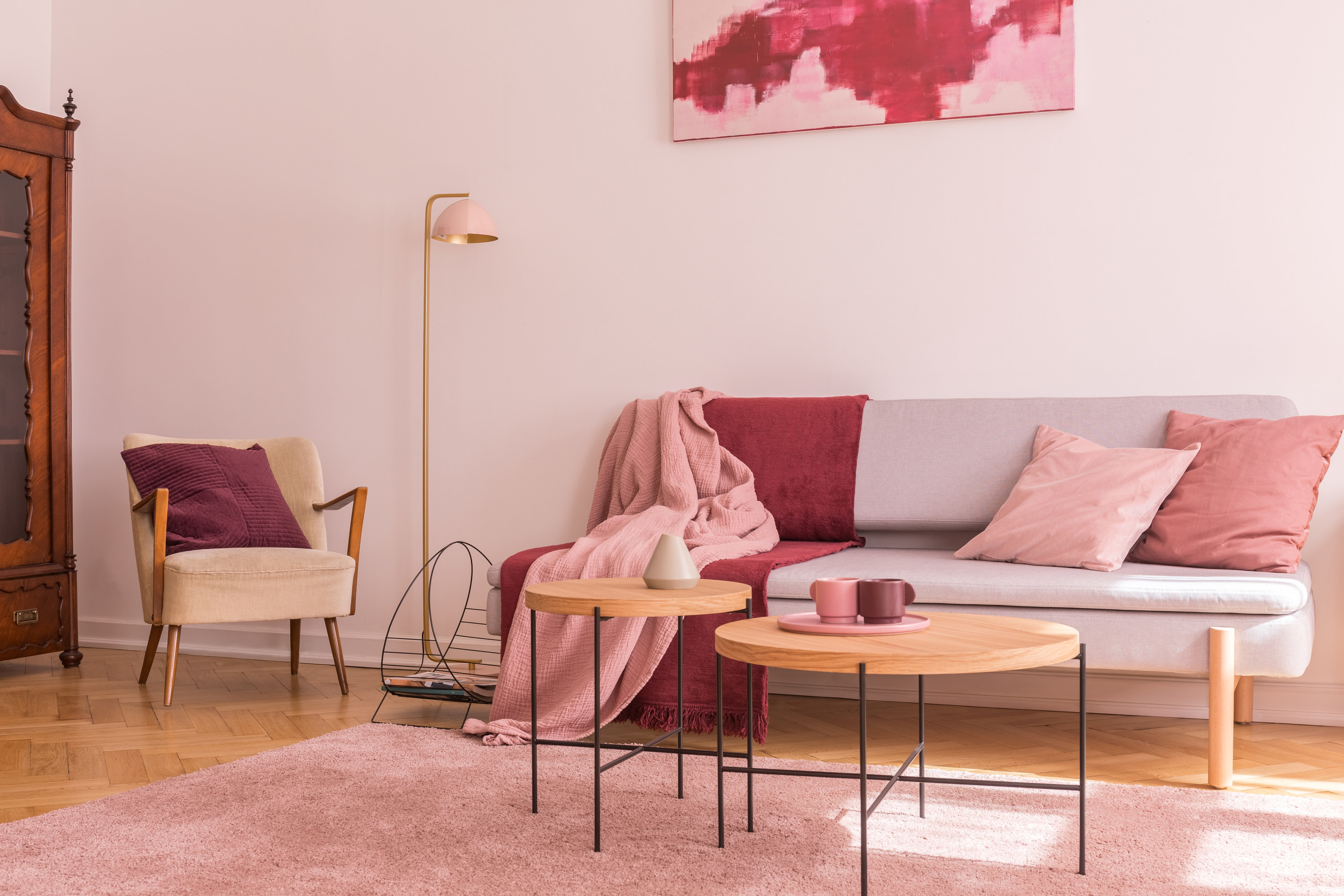 two wooden coffee tables with pink coffee mugs on them next to modern grey sofa with pillows and blankets in different shades of pink, a pink fuzzy rug, a pink lamp, and a pink painting on the wall