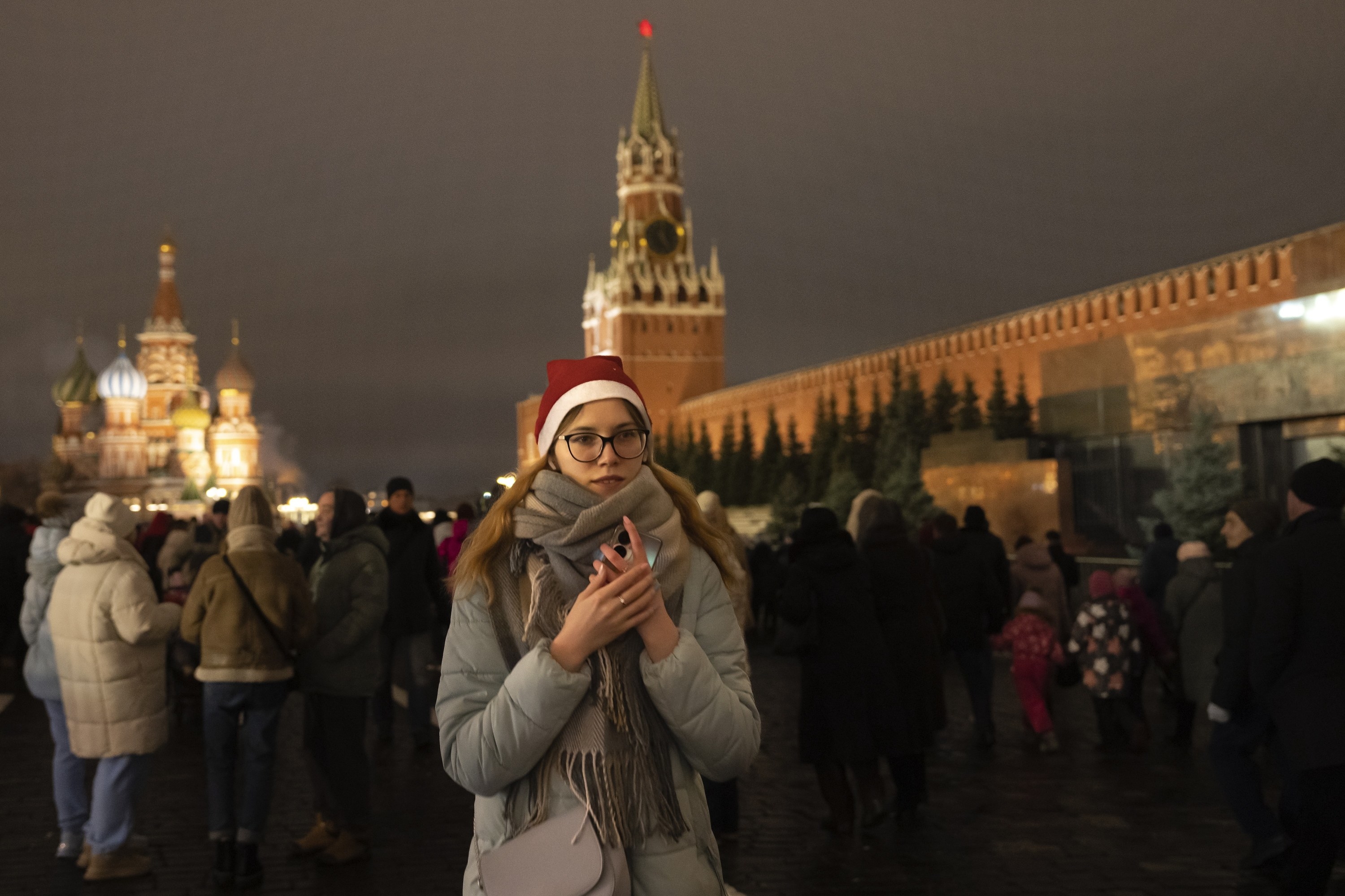 A woman stands in a crowded public square between St. Basil&#x27;s Cathedral to the left and the Spasskaya Tower to the right
