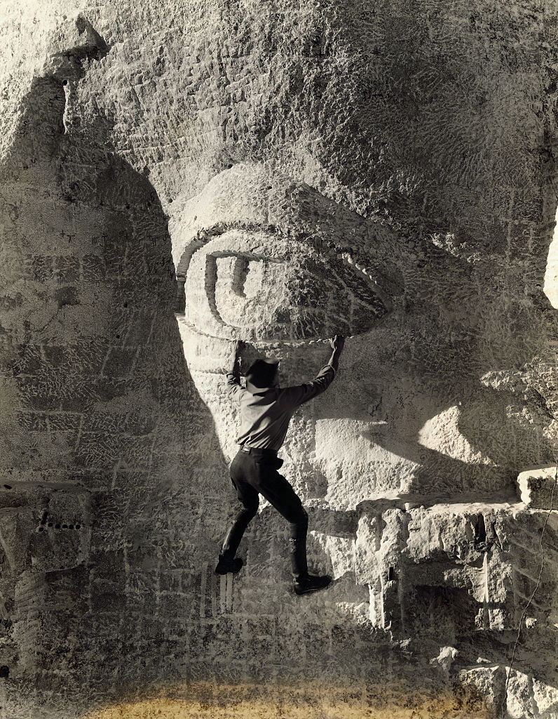 Person wearing a wide-brimmed hat and perched on the side of the mountain and carving the eyes