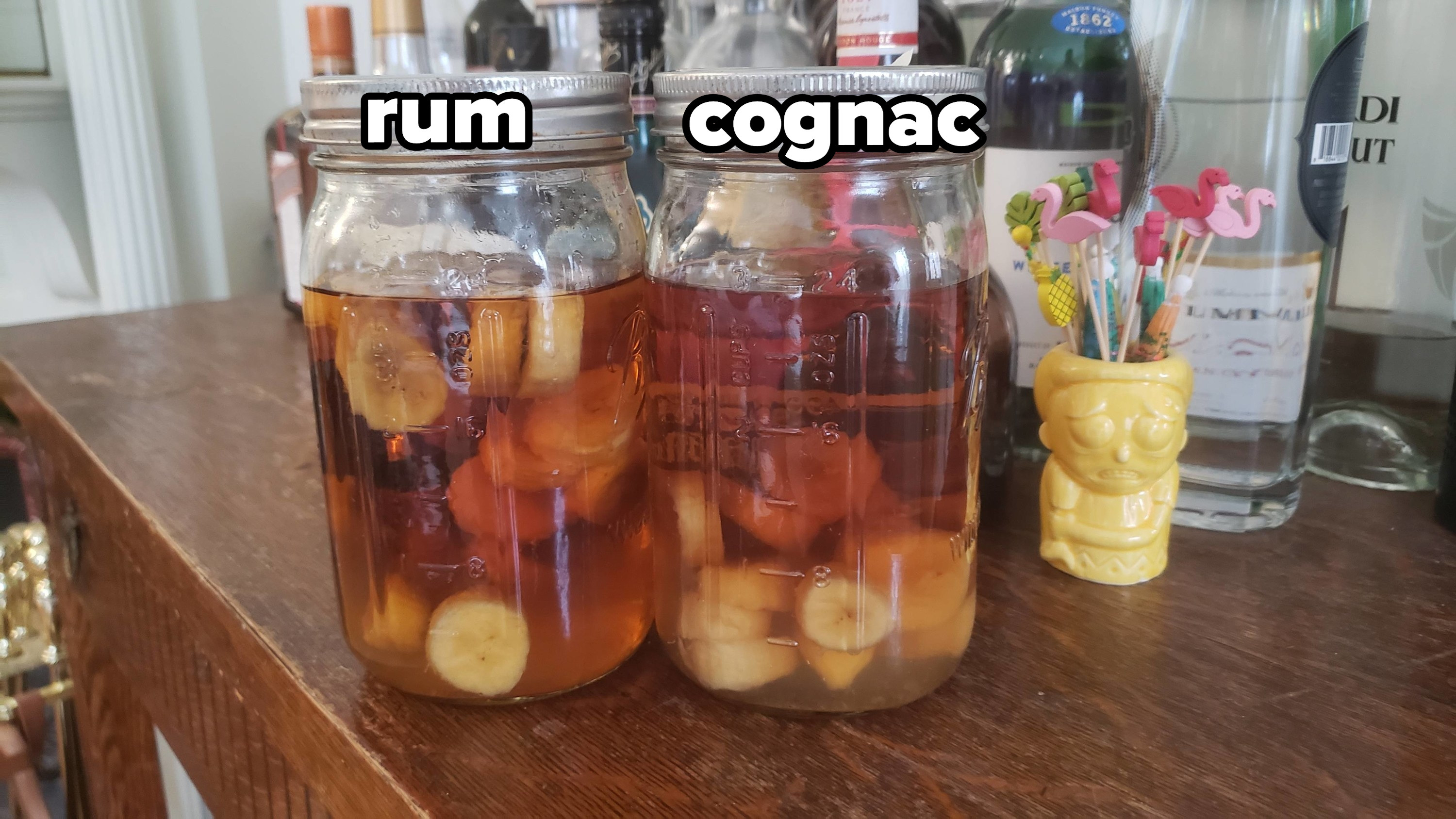 Two glasses, one of rum and one of cognac, with bananas floating in rum and at the bottom of the cognac