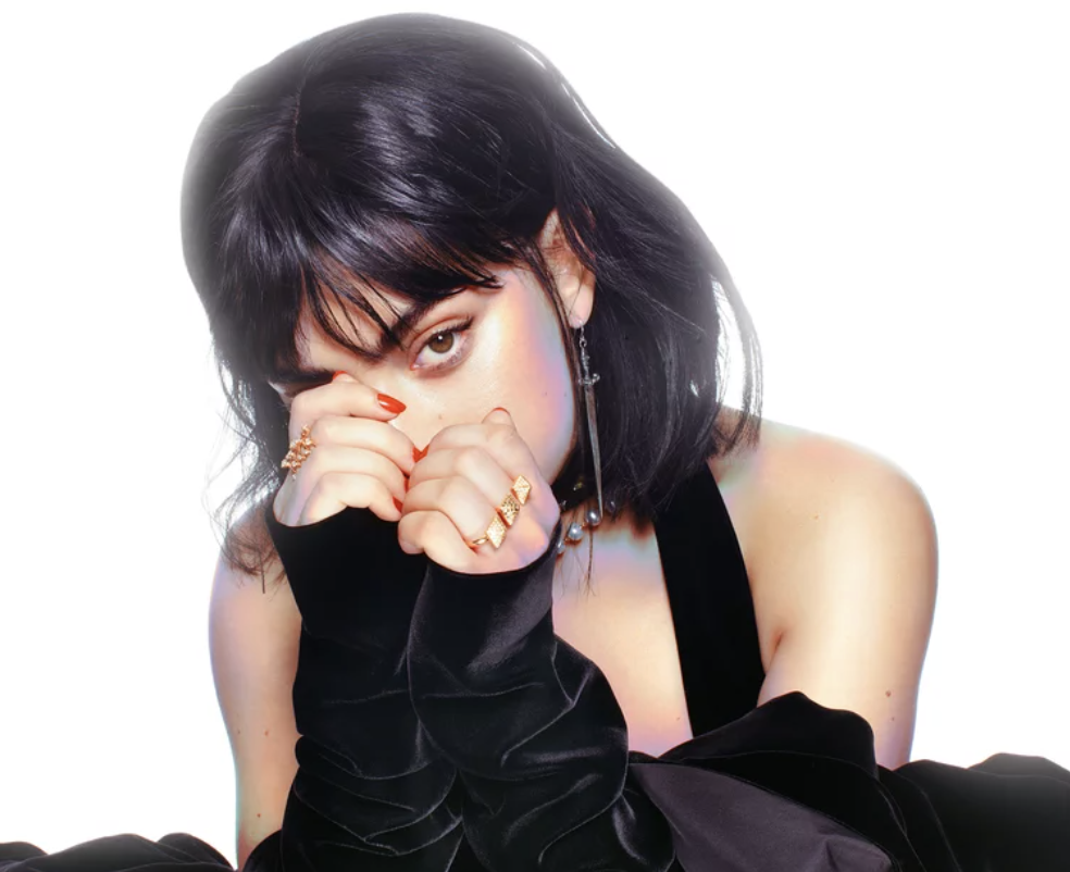 charli wears a tank top and long-sleeved velvet arm warmers. she has a shoulder-length bob cut with bangs and hides behinds her hands
