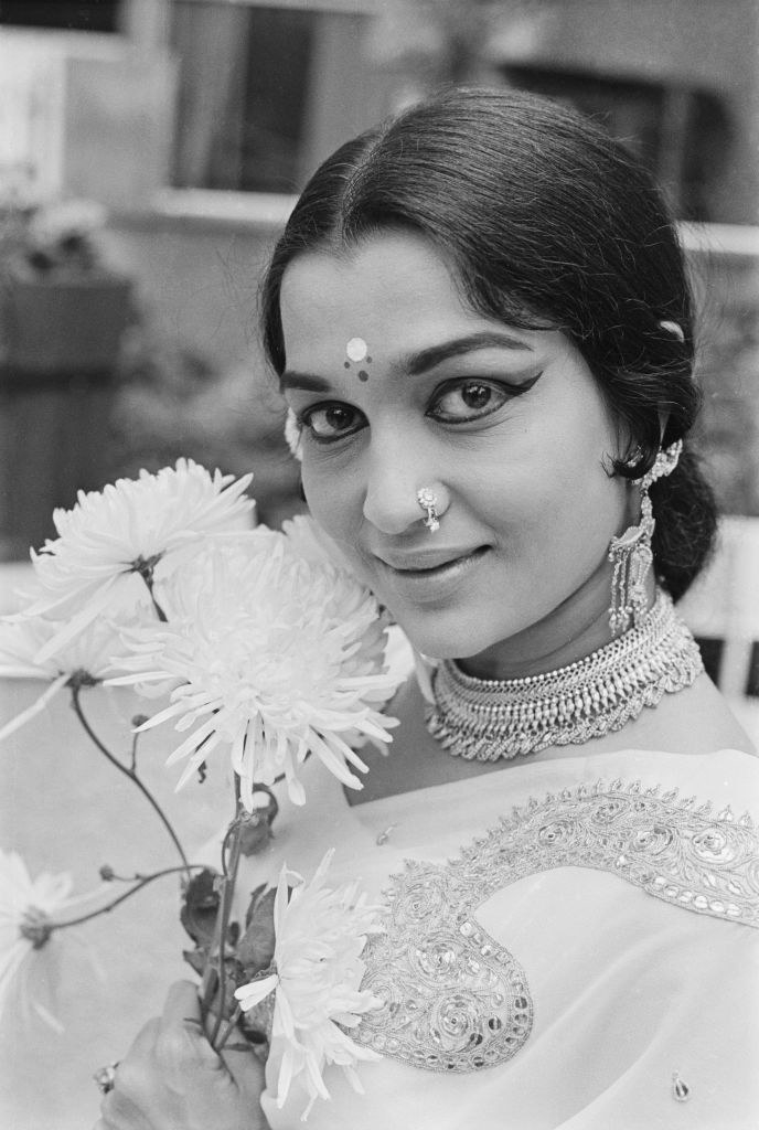 Indian actress Asha Parekh wearing a nose piercing, earrings and a necklace as she poses beside a flower