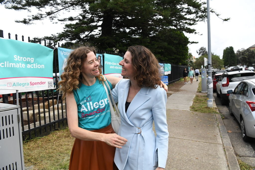 Two women embrace on a sidewalk, smiling widely at each other, one wearing a light blue jacket and the other a teal t-shirt that reads &quot;vote Allegra Spender&quot; with the sleeves rolled up
