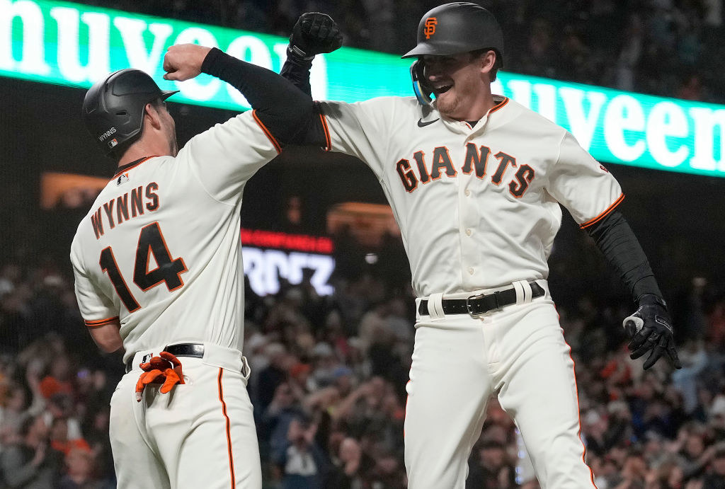 Two baseball players in Giants-branded uniforms and helmets jump in the air and bump elbows in celebration