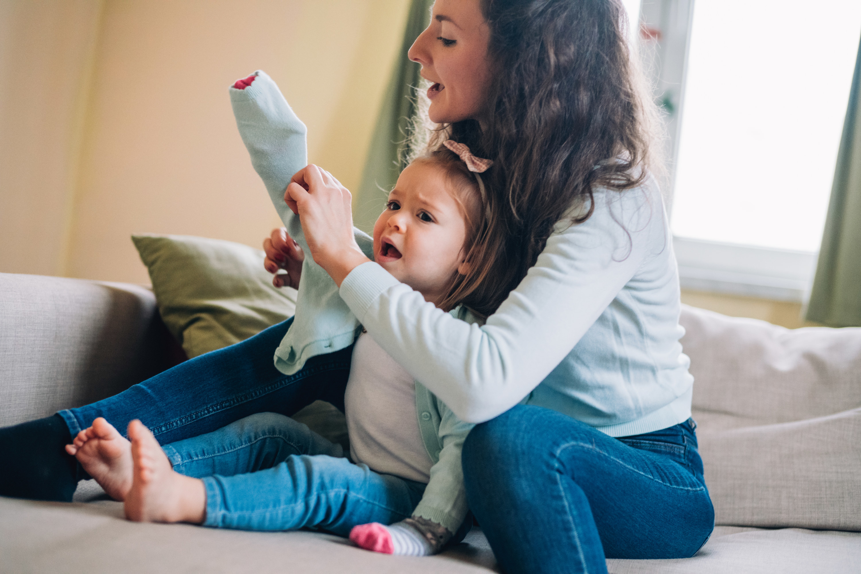 mom sitting behind a toddler trying to put her sweater sleeve on as she cries while wearing a sock on her hand