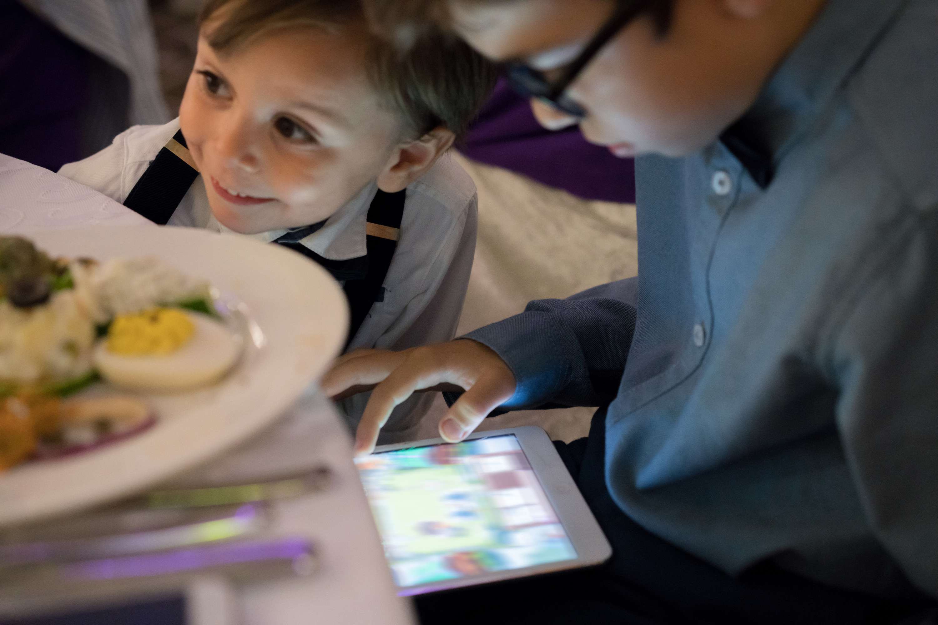 toddler and grade-school aged child dressed in button downs playing with a tablet at a restaurant with a plate of food in front of them