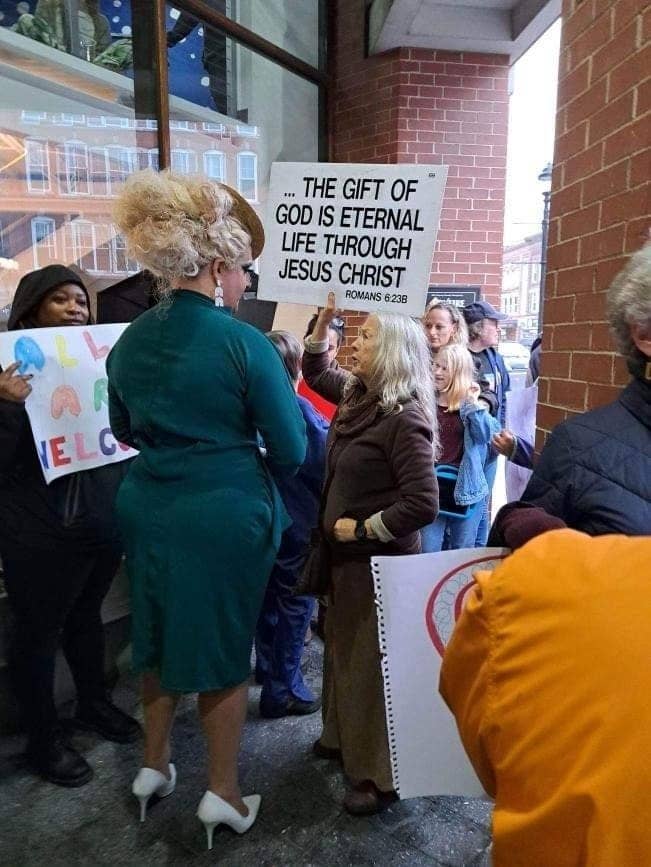 A drag queen wearing white heels and a wig talks to a protester in a group holding a sign reading &quot;the gift of God is eternal life through Jesus Christ&quot;