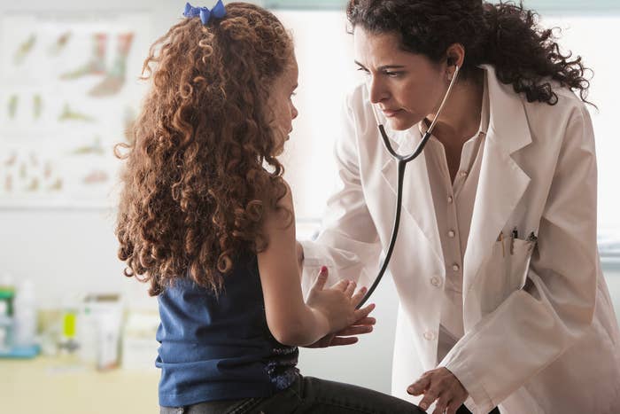 A child sits upright as a doctor wearing a stethoscope leans forward and listens to her heartbeat