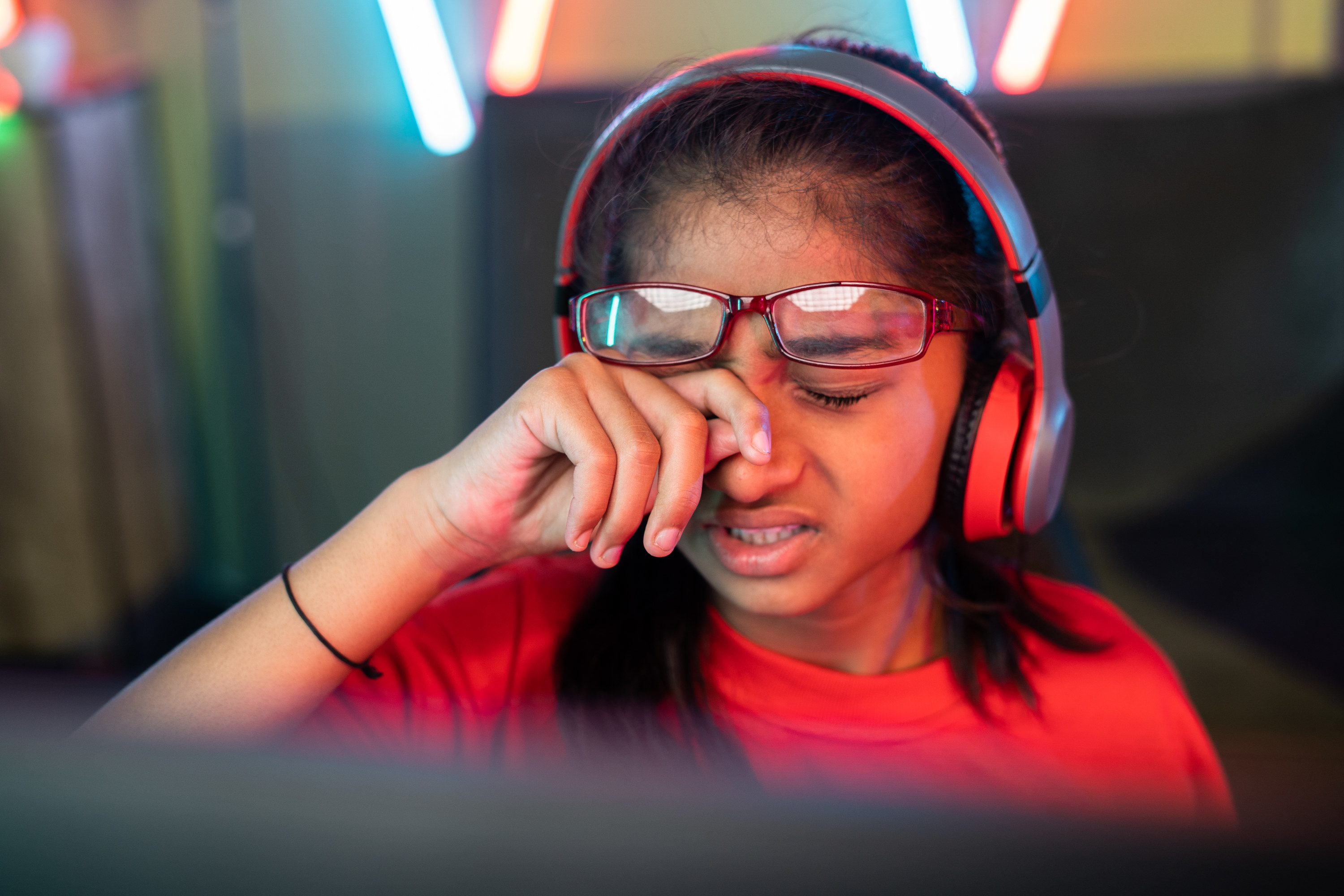 girl rubbing her eyes beneath her glasses w hile wearing headphones and looking tired and frustrated in front of her computer