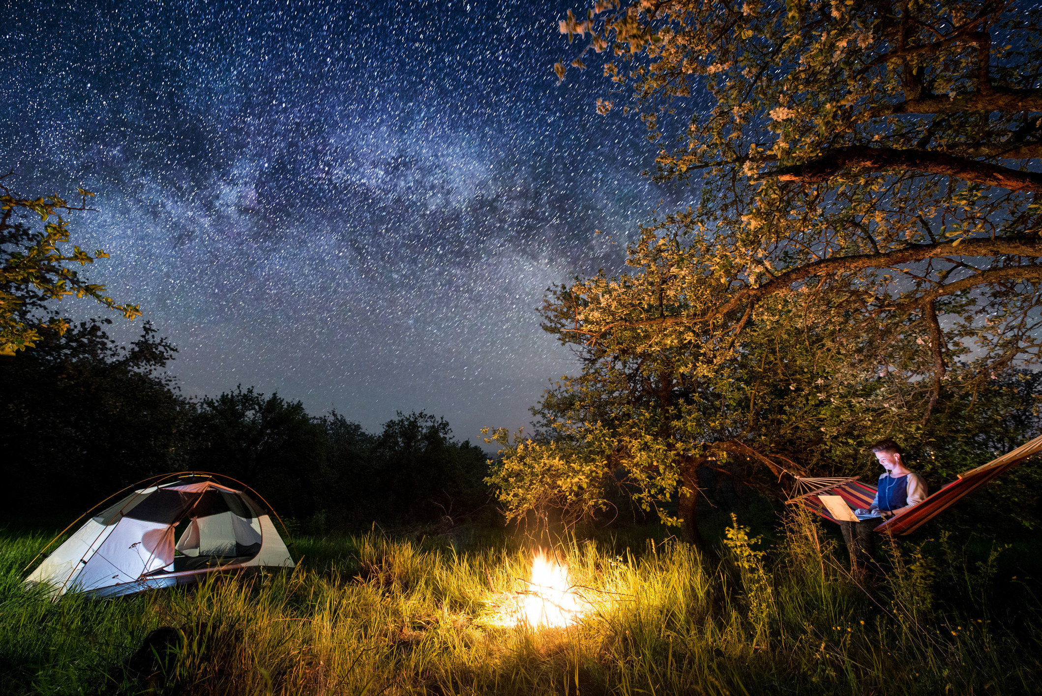 A person sitting in a hammock outdoors at night next to a campfire