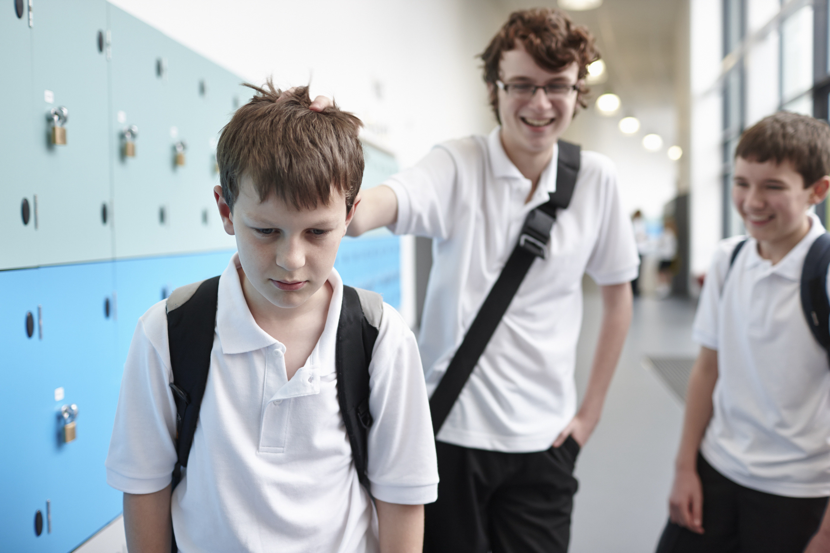 Three boys in a school hallway, with two laughing and one looking upset