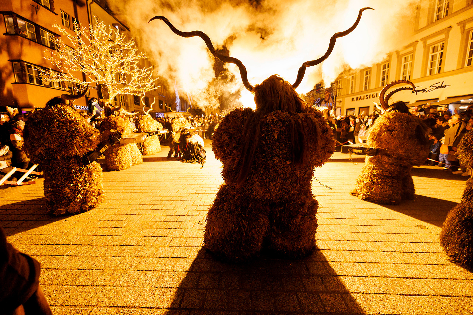 A group of people in heavy robes and long horns look at a yellow smokey fire with their backs turned to the camera