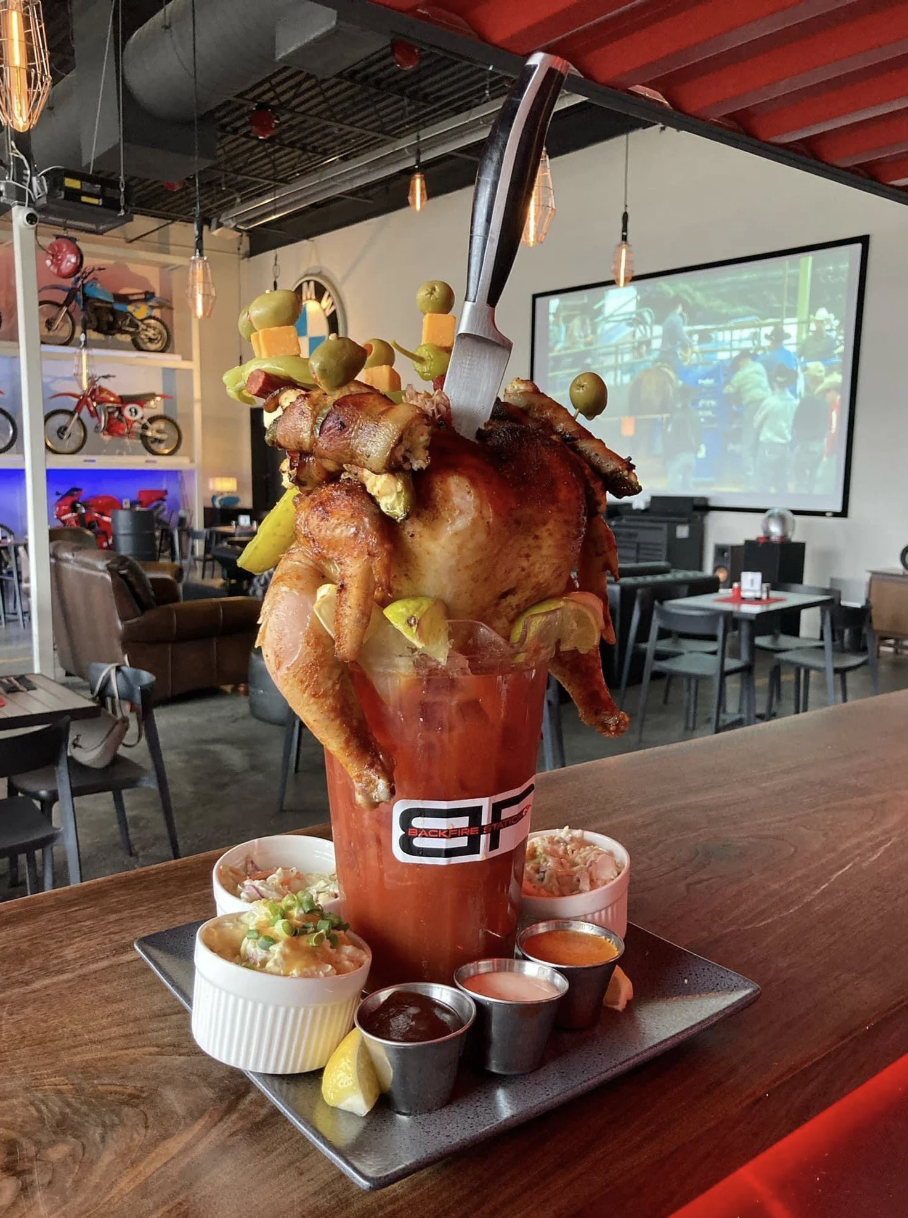 A full chicken with bacon and other condiments shoved inside a cup; there&#x27;s too much food for the cup, so it&#x27;s spilling out the sides
