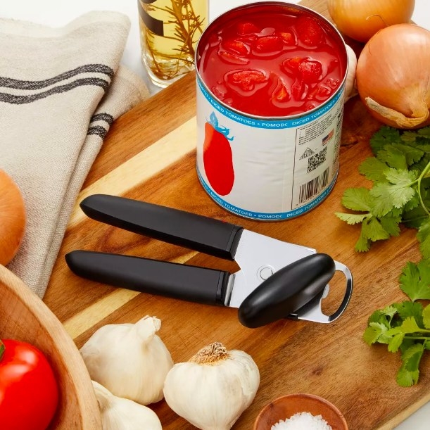 Can opener next to various ingredients and an open can of tomatoes