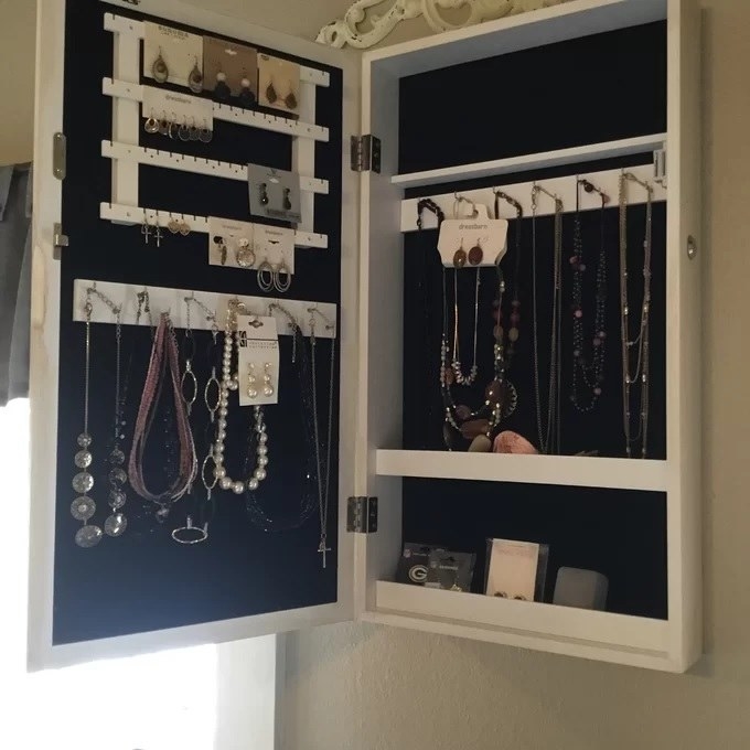 The white jewelry armoire mounted on a wall with a lot of jewelry stored away