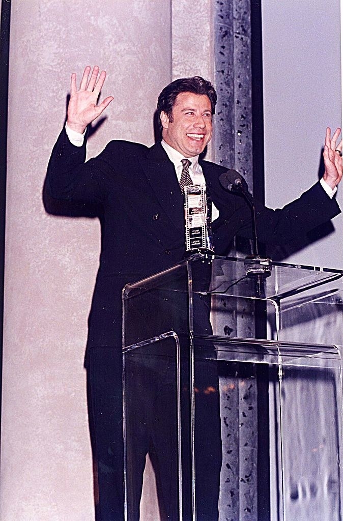 John smiling and holding up his hands at a podium