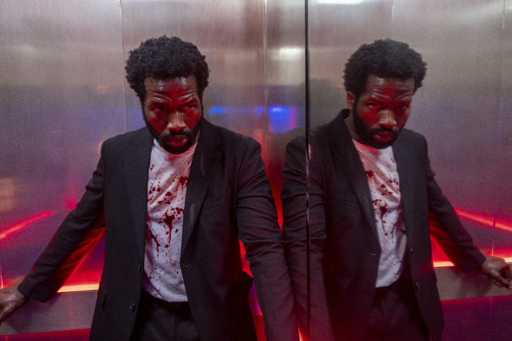 A tired and bloodied man looks at his reflection on the walls of an elevator