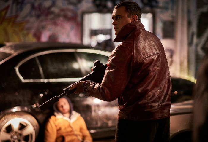 A young man in a leather jacket and a rifle observes the damage of a shootout