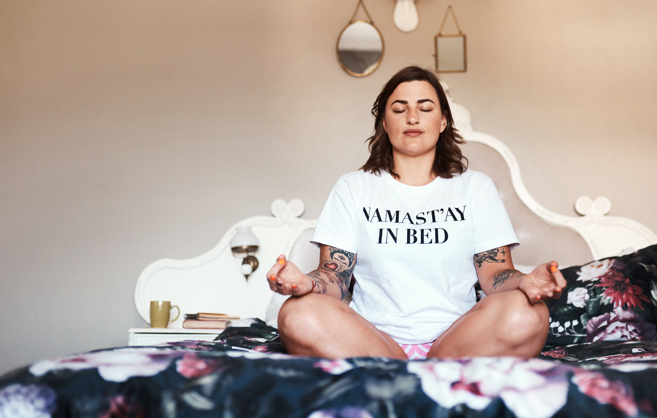 A girl meditates in bed wearing a shirt that says &quot;namastay in bed&quot;