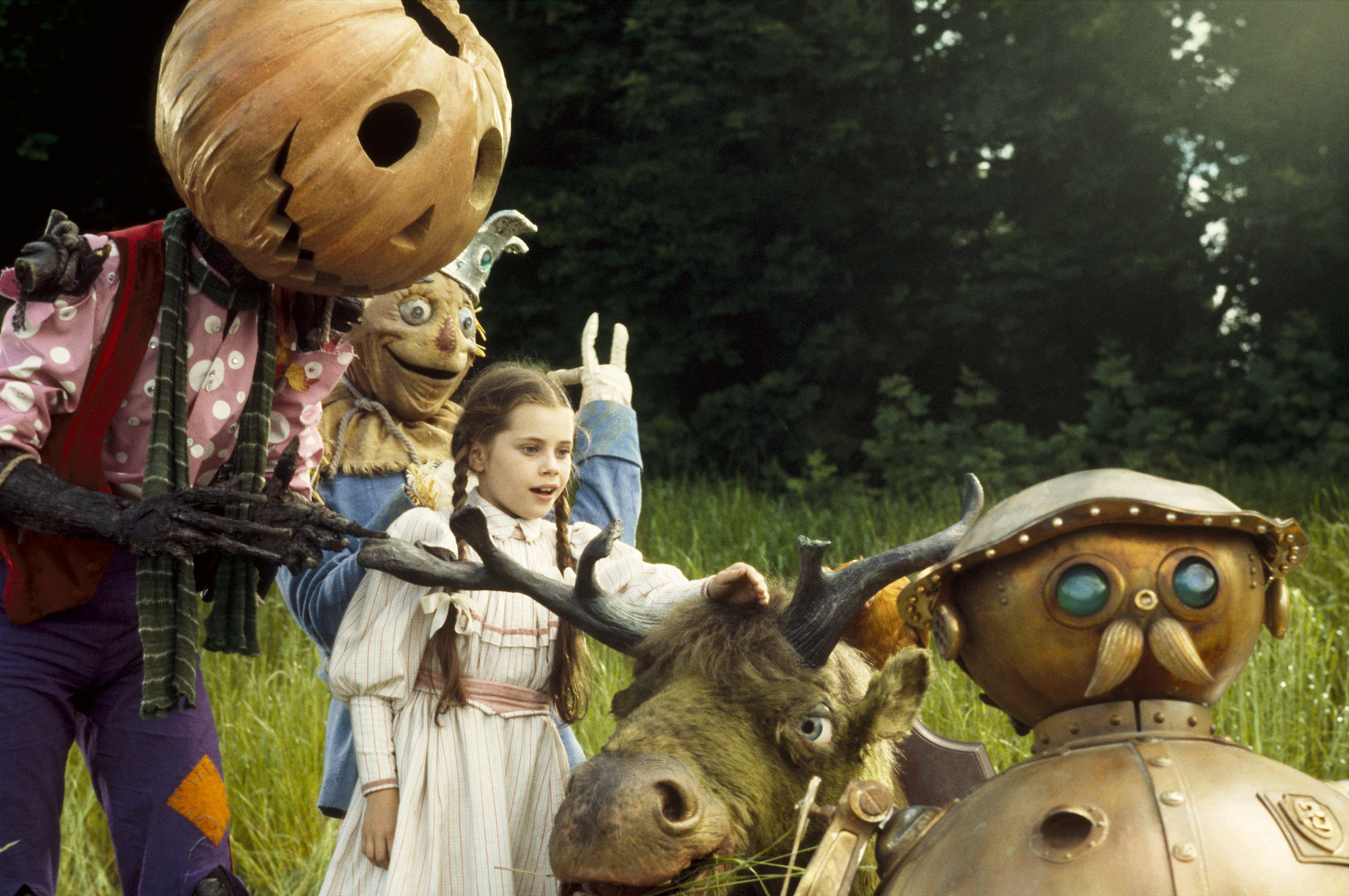 A young girl stands with a scarecrow, a pumpkin man, and a robot