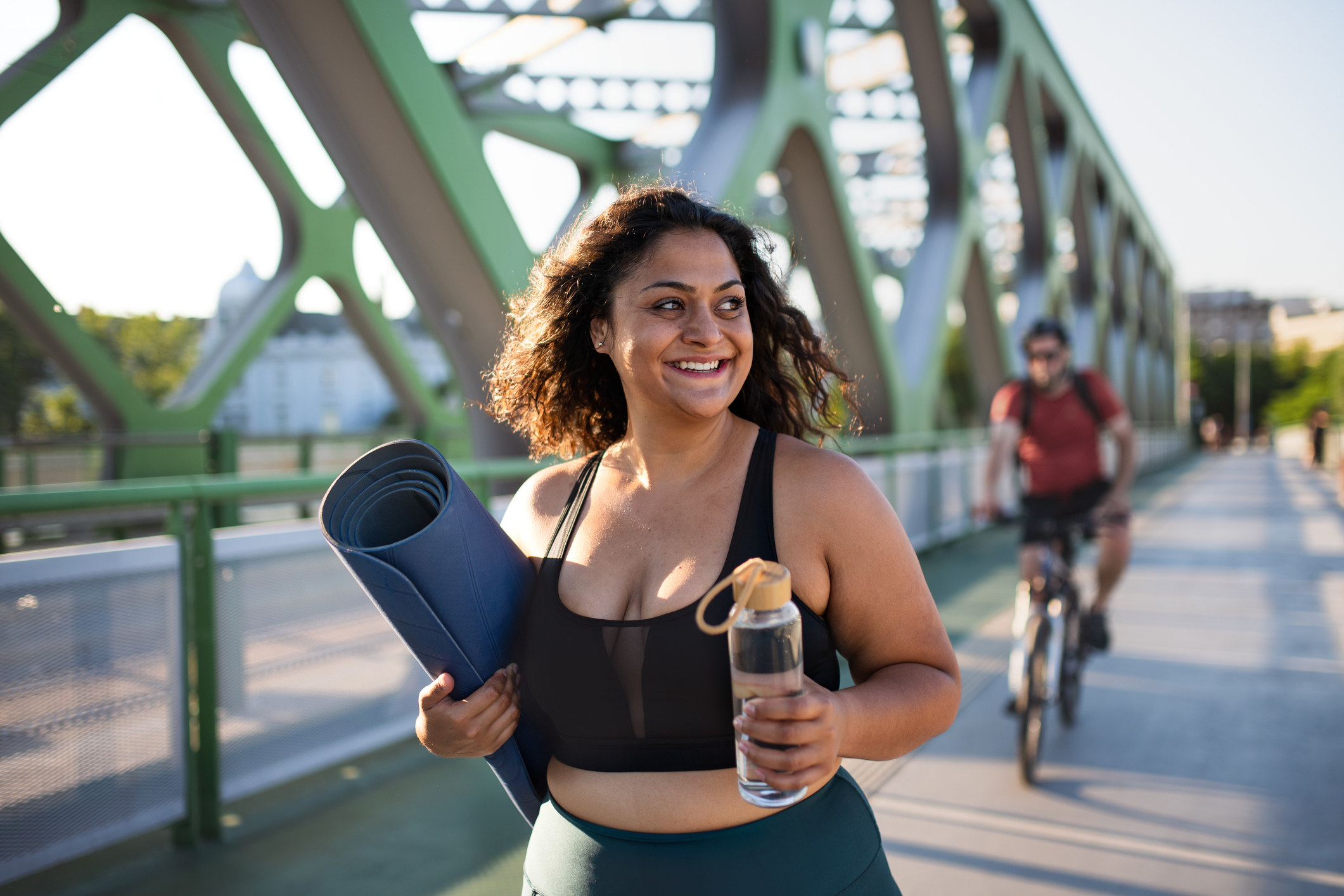 A woman walks home from a workout class with a yoga mat and a water bottle