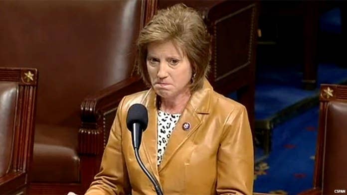 A representative wears a beige leather jacket, standing on the floor of the House of Representatives in front of a microphone and frowning