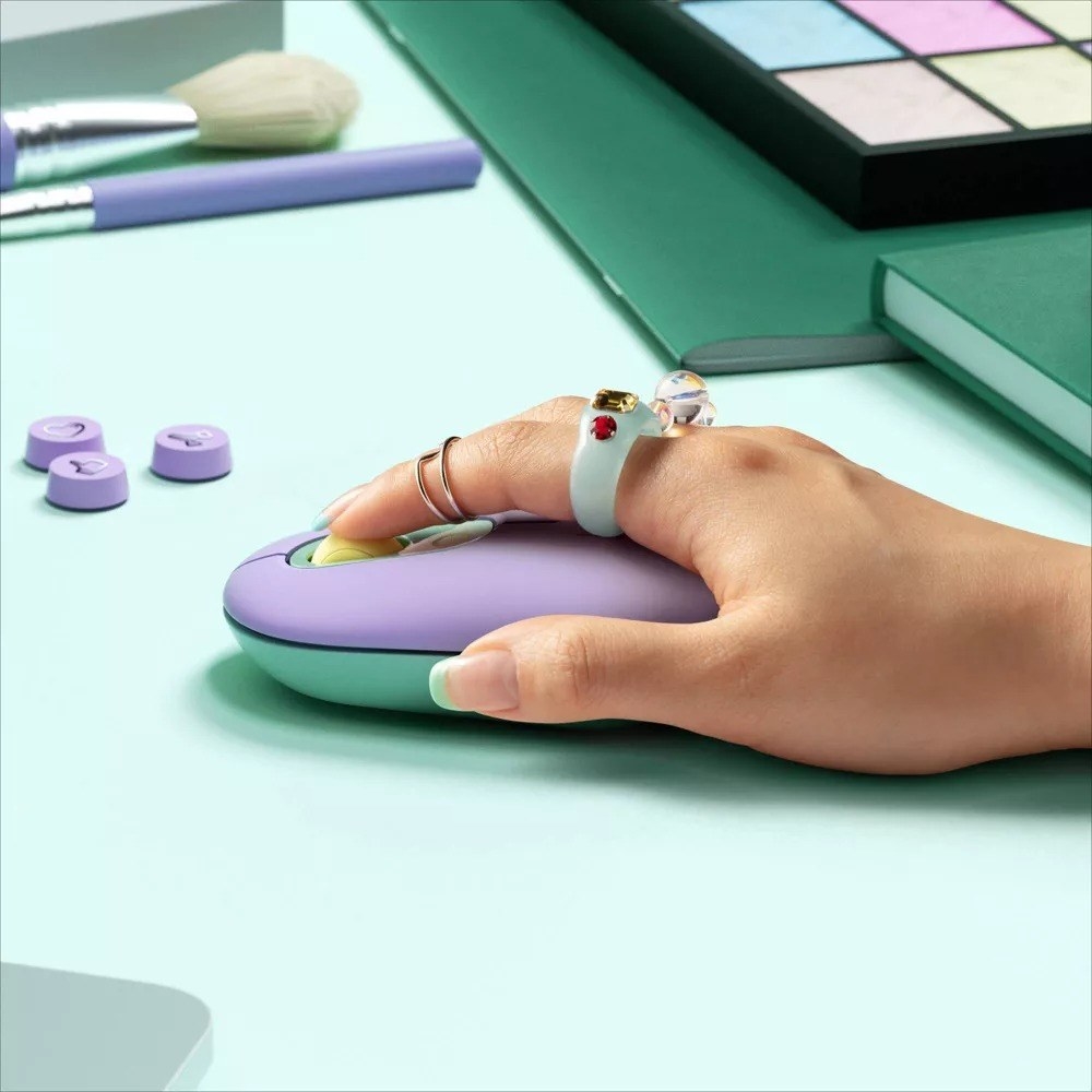 A model&#x27;s hand using the lavender mouse over a teal surface with lavender computer keys and makeup strewn around.