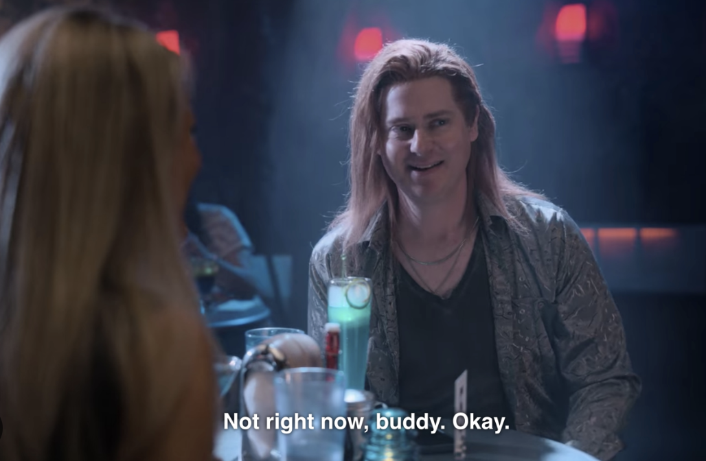 a man with long hair down to his shoulders wears a v-neck and linen shirt while sitting at a restaurant table. he says to someone off-camera, "not right now buddy, okay."