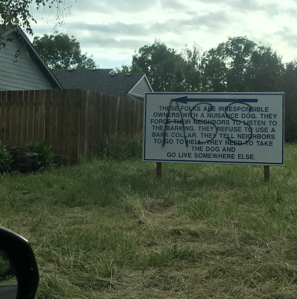 A large yard sign that says the people living next door are bad owners of a nuisance dog; the sign has been spray-painted with the word &quot;ass&quot; over the message