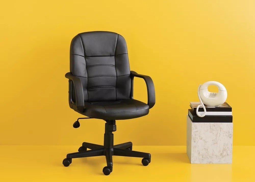 The black ergonomic office chair against the yellow background next to an art deco style phone and side table