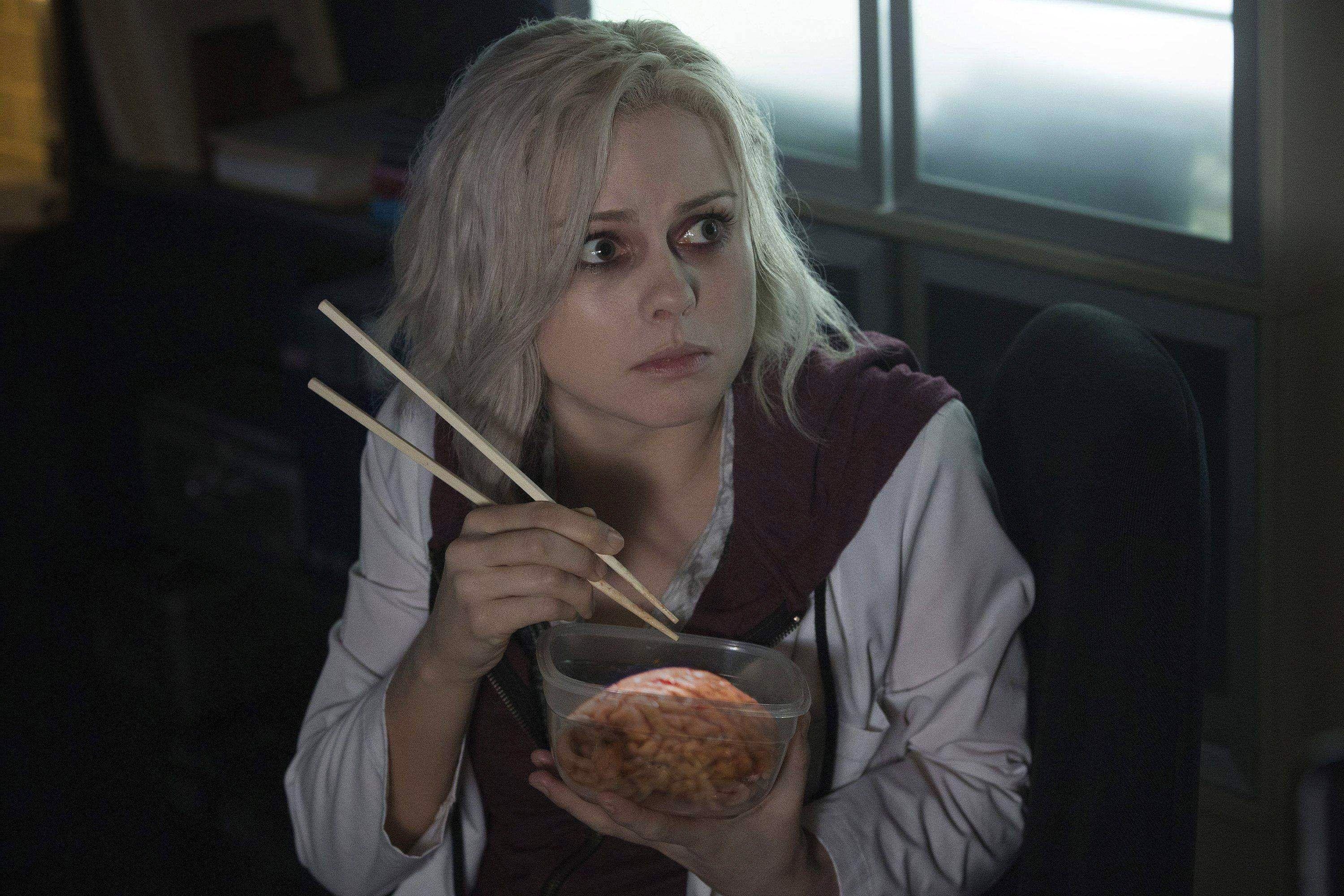 A pale blonde woman eats a brain out of tupperware with chopsticks