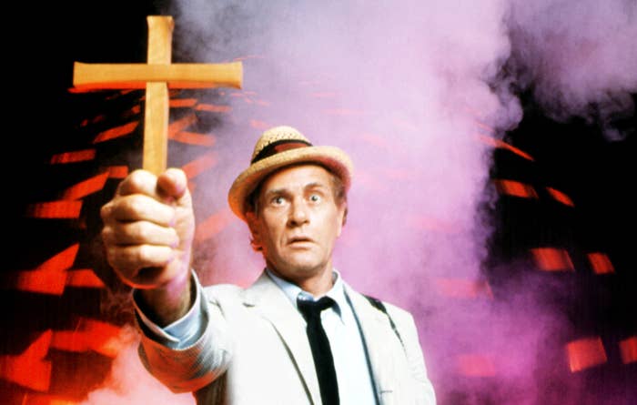 A middle-aged man in a white suit and hat holds a cross against a foggy and surreal background