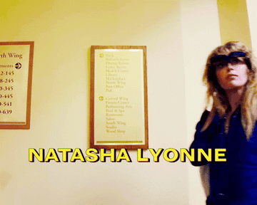 Natasha Lyonne walks down a hallway with flip sunglasses and a blue jacket