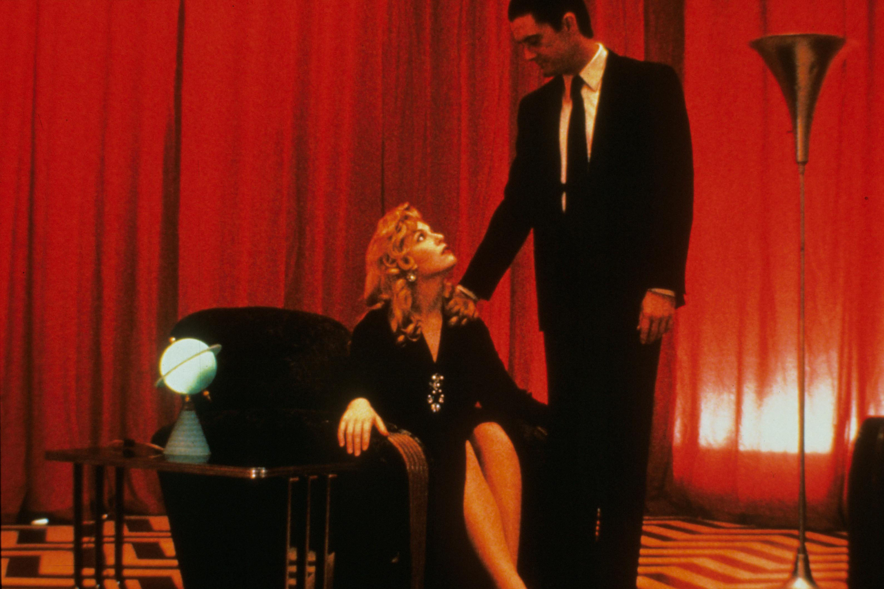 A man in a three-piece suit stands in a red draped room next to a blonde woman on a leather chair