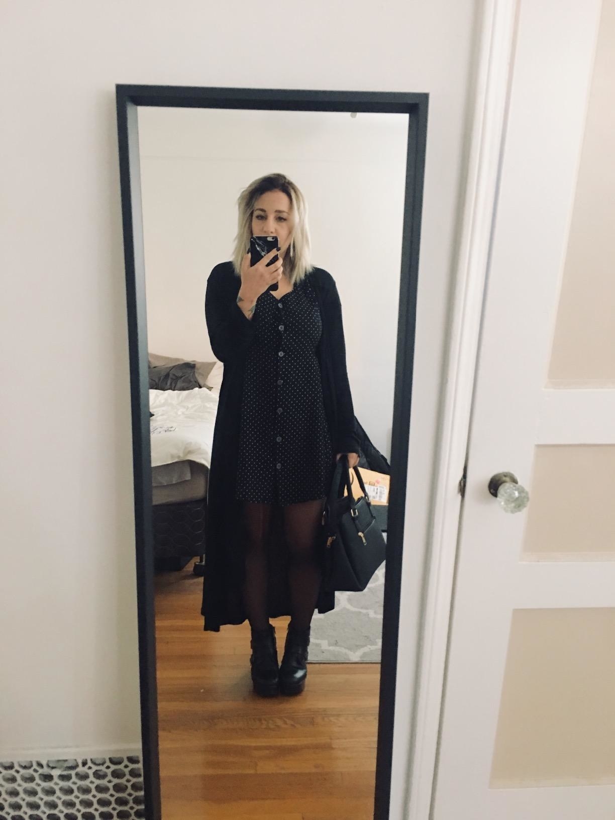 Reviewer wearing long black cardigan over black dress with black tights and boots