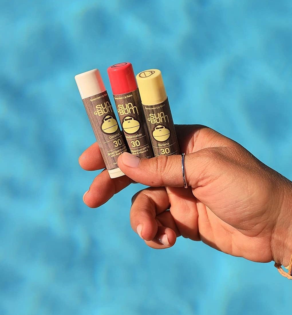 a person holding three tubes of lip balm over vibrant water
