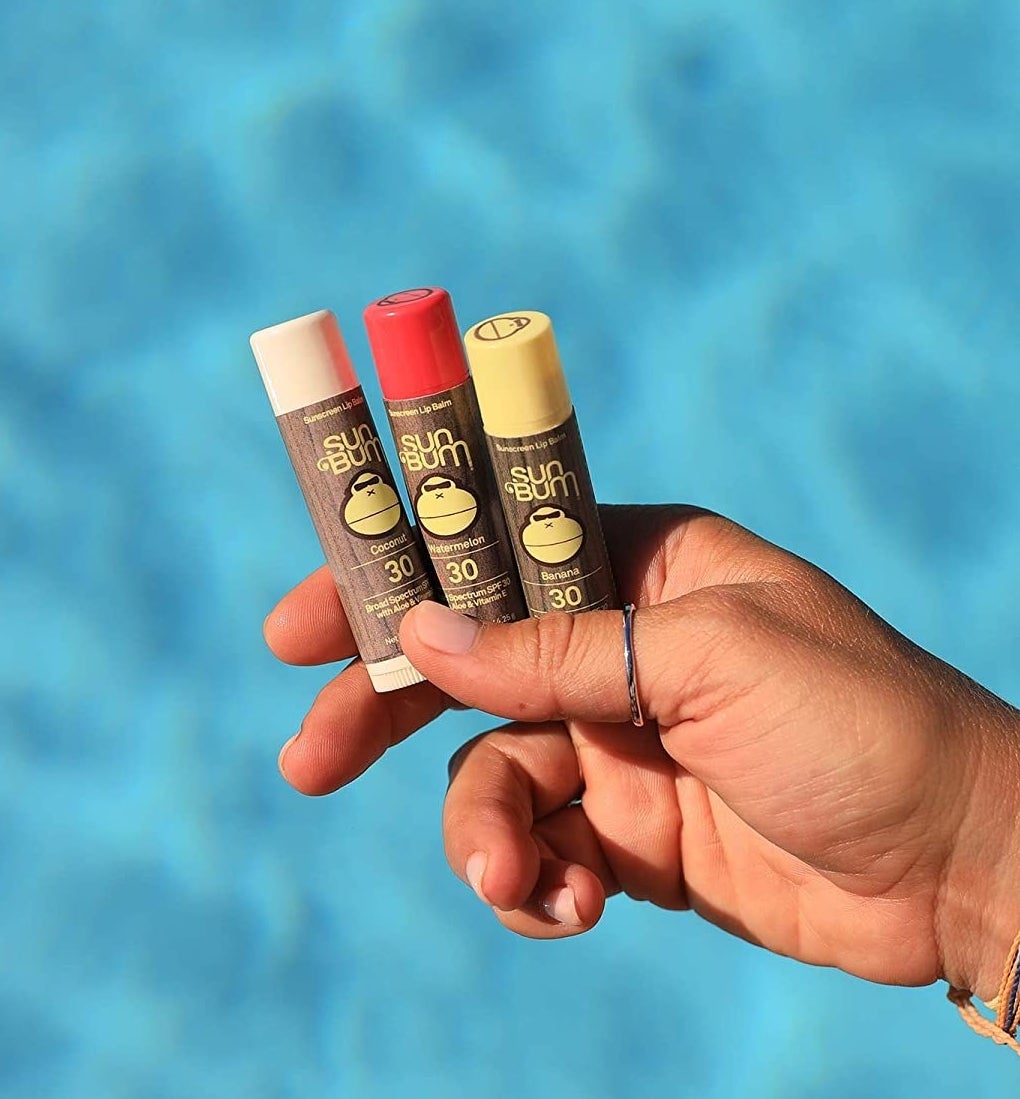 a person holding three tubes of lip balm over vibrant water