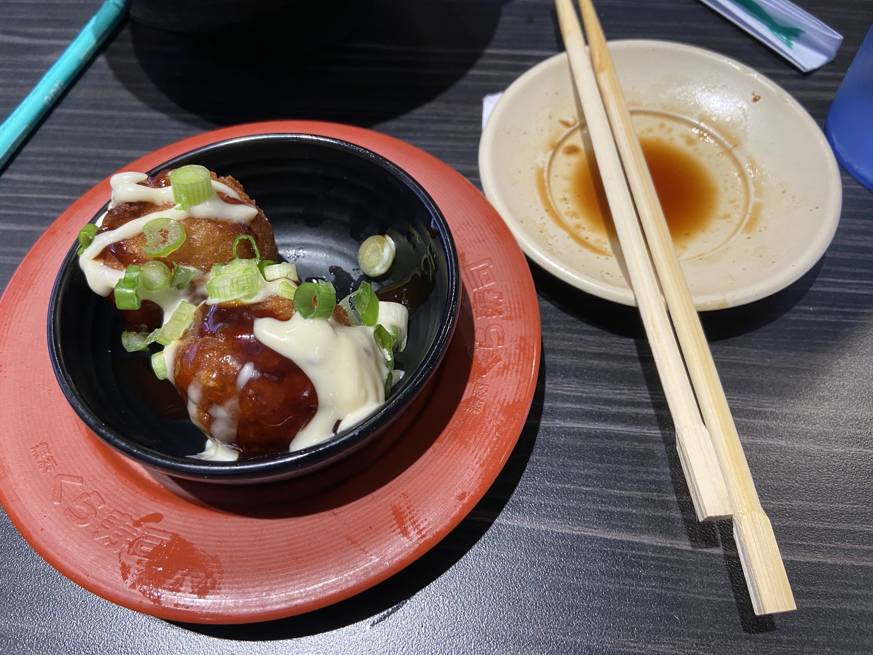 A small plate of takoyaki alonside chopsticks resting on top of a soy sauce dish